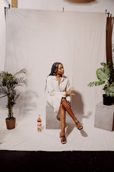 A seated person on a block holds a wine glass, surrounded by potted plants. They wear a light-colored dress and sandals. A bottle of wine is on the floor nearby. The setting is minimalist with a light backdrop.