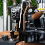 A coffee station featuring a shiny espresso machine with wooden handles and stainless steel details. A black coffee grinder with the brand name engraved on it stands nearby, accompanied by a tamper and a container on a wooden countertop. The setup is elegant and modern with some green plants blurred in the background.