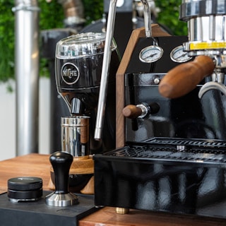 A coffee station featuring a shiny espresso machine with wooden handles and stainless steel details. A black coffee grinder with the brand name engraved on it stands nearby, accompanied by a tamper and a container on a wooden countertop. The setup is elegant and modern with some green plants blurred in the background.