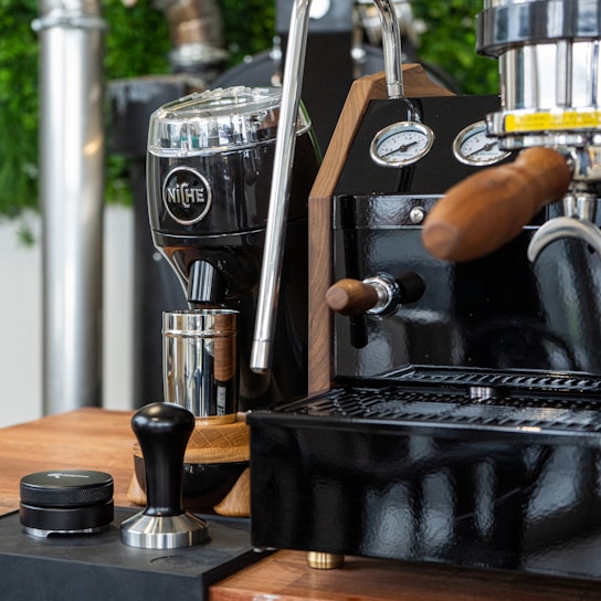 A coffee station featuring a shiny espresso machine with wooden handles and stainless steel details. A black coffee grinder with the brand name engraved on it stands nearby, accompanied by a tamper and a container on a wooden countertop. The setup is elegant and modern with some green plants blurred in the background.