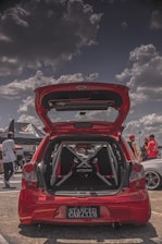 A red hatchback car is parked with its trunk open, showcasing a custom interior with a visible roll cage and 'Etios' branding. The scene is set in what appears to be a car show with partly cloudy skies. Several people are gathered around, some dressed in casual attire, wearing hats and sunglasses. There is a tent with 'Volcano' branding in the background.