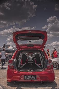 A red hatchback car is parked with its trunk open, showcasing a custom interior with a visible roll cage and 'Etios' branding. The scene is set in what appears to be a car show with partly cloudy skies. Several people are gathered around, some dressed in casual attire, wearing hats and sunglasses. There is a tent with 'Volcano' branding in the background.