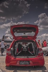 A red hatchback car is parked with its trunk open, showcasing a custom interior with a visible roll cage and 'Etios' branding. The scene is set in what appears to be a car show with partly cloudy skies. Several people are gathered around, some dressed in casual attire, wearing hats and sunglasses. There is a tent with 'Volcano' branding in the background.