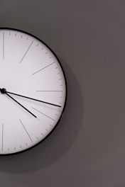 A minimalistic white wall clock with thin black hands on a plain wall.