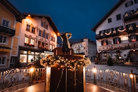 A charming town square at night, adorned with festive lights and decorations. Traditional buildings with ornate facades and balconies line the square, featuring shops and a restaurant. A central monument is decorated with garlands and lights, creating a warm, inviting atmosphere.