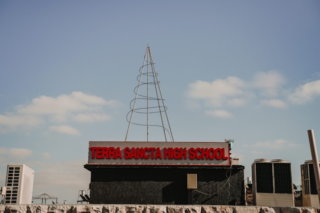 A high school sign with the name 'Terra Sancta High School' in prominent red letters, set against a blue sky with scattered clouds. There is a wireframe structure resembling a tree or antenna on top of the sign. Air conditioning units and some industrial elements are visible in the background.