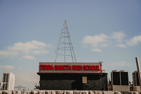 A high school sign with the name 'Terra Sancta High School' in prominent red letters, set against a blue sky with scattered clouds. There is a wireframe structure resembling a tree or antenna on top of the sign. Air conditioning units and some industrial elements are visible in the background.