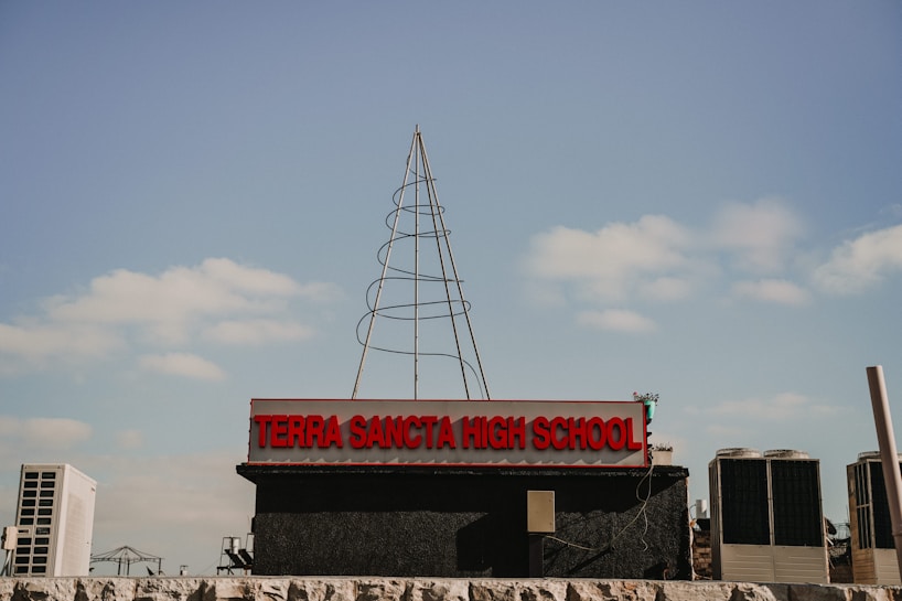 A high school sign with the name 'Terra Sancta High School' in prominent red letters, set against a blue sky with scattered clouds. There is a wireframe structure resembling a tree or antenna on top of the sign. Air conditioning units and some industrial elements are visible in the background.