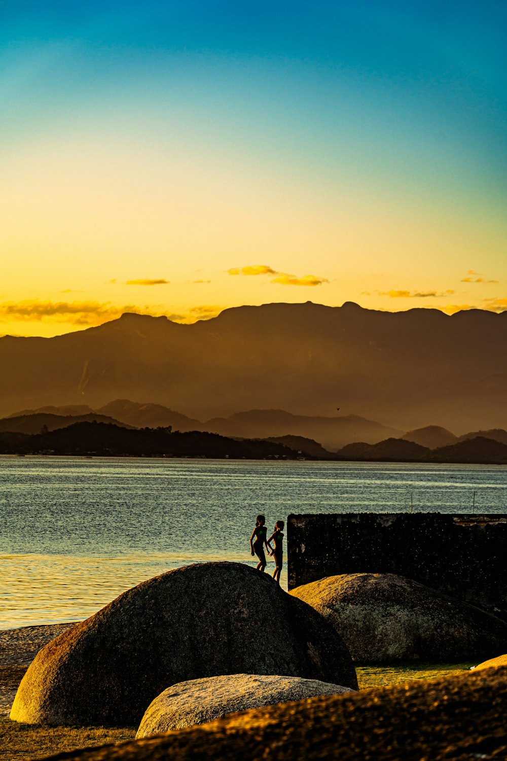 silhouette of 2 people standing on rock formation near body of water during sunset