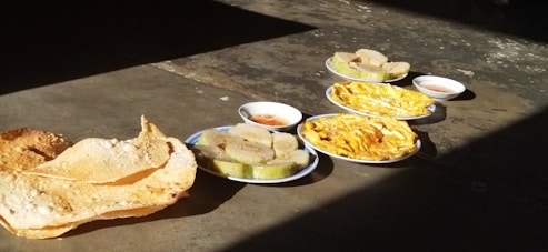 A variety of dishes is spread out on a concrete surface in bright sunlight. The meal includes large crisp papadums, slices of a loaf cake, possibly layered with filling, and what appears to be omelet or scrambled eggs served on oval plates. Small bowls of dipping sauce accompany the dishes.