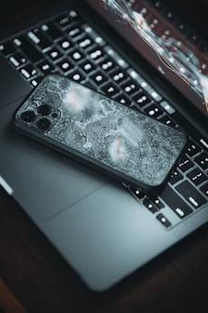 A smartphone with a decorative case lies on top of an open laptop, with the keyboard and a segment of the screen visible. The laptop's screen is displaying a futuristic, abstract interface of charts and curves. The setting has a moody ambience with dim lighting.