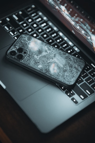 A smartphone with a decorative case lies on top of an open laptop, with the keyboard and a segment of the screen visible. The laptop's screen is displaying a futuristic, abstract interface of charts and curves. The setting has a moody ambience with dim lighting.