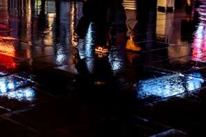 Stylish leather boots stepping on wet pavement reflecting neon lights in shades of pink and blue.