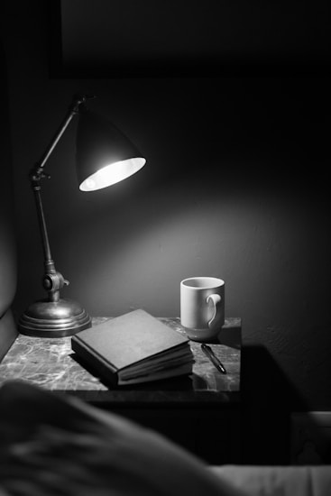 A dimly lit scene features a vintage desk lamp casting soft light over a small table. On the table, there is a closed book next to a ceramic mug and a pen, creating an atmosphere of solitude and contemplation.