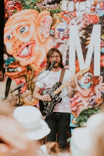 A musician passionately plays an electric guitar on stage in front of a colorful and abstract mural featuring a large caricature face. The setting appears to be outdoors, possibly at a music festival, as indicated by the crowd visible at the bottom of the image.