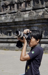 A person is holding a professional camera with a large lens, appearing focused while capturing photographs. The background shows an ancient stone wall with intricate carvings and architectural details.