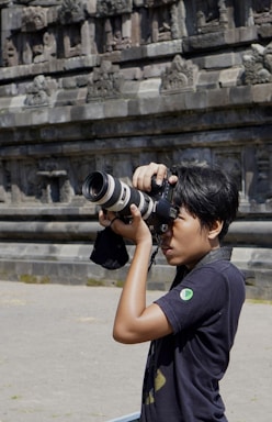 A person is holding a professional camera with a large lens, appearing focused while capturing photographs. The background shows an ancient stone wall with intricate carvings and architectural details.