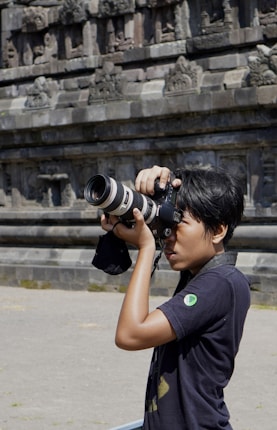 A person is holding a professional camera with a large lens, appearing focused while capturing photographs. The background shows an ancient stone wall with intricate carvings and architectural details.