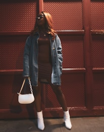 A person wearing a brown dress and denim jacket stands confidently against a textured, dark red background. They sport white ankle boots and hold a small white purse. Their hair is long and styled, and they wear sunglasses.