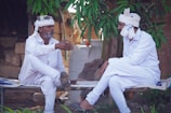 Two men dressed in white traditional attire are sitting on a woven cot. They are engaged in conversation under a leafy tree, with one man gesturing with his hand while the other listens attentively. The background features rustic elements, including bricks and a water container.