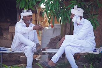 Two men dressed in white traditional attire are sitting on a woven cot. They are engaged in conversation under a leafy tree, with one man gesturing with his hand while the other listens attentively. The background features rustic elements, including bricks and a water container.