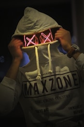 A person is wearing a light gray hoodie with 'MAXZONE' printed on it. They are holding the hood over their face, revealing a pair of glowing, cross-stitched, X-shaped eyes that give a mysterious and intriguing appearance.