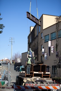 Construction workers are securing a large steel beam being lifted by a crane, positioned next to a building. The workers are wearing safety gear including helmets and reflective vests. The scene is set outdoors with clear skies and modern buildings in the background.