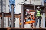 Construction workers are on site assembling a steel structure. One worker is on an orange lift, securing or inspecting a beam, while another stands nearby wearing a safety vest and helmet.