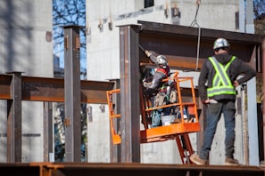 Construction workers are on site assembling a steel structure. One worker is on an orange lift, securing or inspecting a beam, while another stands nearby wearing a safety vest and helmet.