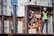 Construction workers are on site assembling a steel structure. One worker is on an orange lift, securing or inspecting a beam, while another stands nearby wearing a safety vest and helmet.