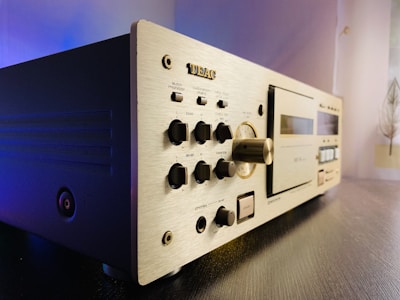 A vintage audio cassette deck with a brushed aluminum front panel featuring various knobs and buttons for adjusting settings such as bias, level, and balance. On the right side, there is a tape compartment with a clear window, and control switches below. The deck is placed on a dark wooden surface, with soft lighting creating a subtle blue and yellow gradient background.