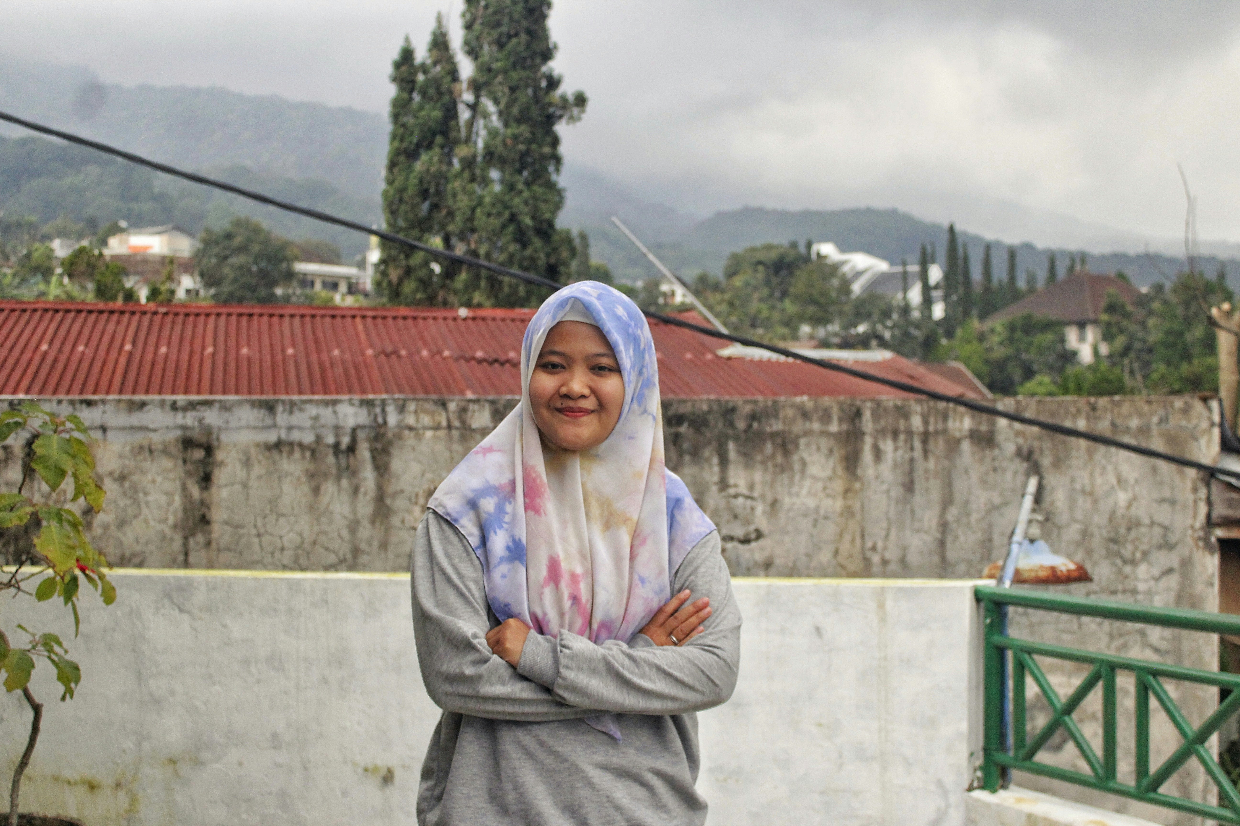 Wanita berjilbab abu-abu berdiri di dekat pagar kayu coklat