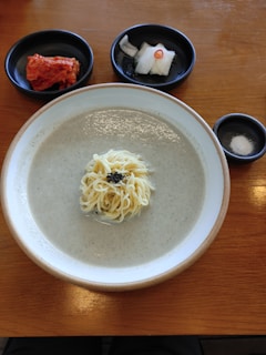 A bowl of creamy soup with noodles is centered on a wooden table. To the side, there are small black dishes containing kimchi and pickled radish slices with a slice of red chili. Another small dish contains salt.