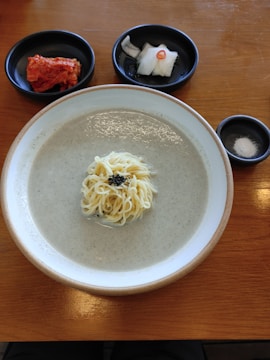 A bowl of creamy soup with noodles is centered on a wooden table. To the side, there are small black dishes containing kimchi and pickled radish slices with a slice of red chili. Another small dish contains salt.