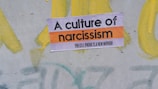 A sign with the text 'A culture of narcissism' and 'the cellphone is a new mirror' in bold letters is placed on a painted surface with swirls of yellow and hints of green and gray in the background.