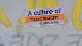 A sign with the text 'A culture of narcissism' and 'the cellphone is a new mirror' in bold letters is placed on a painted surface with swirls of yellow and hints of green and gray in the background.