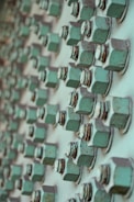 A close-up view of multiple rusted green bolts and nuts attached to a metallic surface, creating a geometric pattern of repeating elements. The bolts show signs of wear and oxidation, contributing to a textured and industrial appearance.