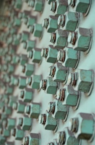A close-up view of multiple rusted green bolts and nuts attached to a metallic surface, creating a geometric pattern of repeating elements. The bolts show signs of wear and oxidation, contributing to a textured and industrial appearance.