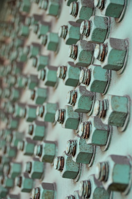 A close-up view of multiple rusted green bolts and nuts attached to a metallic surface, creating a geometric pattern of repeating elements. The bolts show signs of wear and oxidation, contributing to a textured and industrial appearance.