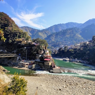 A small village is nestled in a mountainous region, with colorful buildings situated along the banks of a flowing river. The river has a striking green-blue hue, and a few stairs lead down to its edge. A metal bridge spans the river, connecting different parts of the village. Lush greenery and dense forested areas cover the hills, while the sky above is clear with a few wispy clouds.