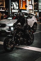 A person wearing a black leather jacket and helmet rides a motorcycle in a city street at night. Bright storefront lights illuminate the background, and a white car is parked nearby, enhancing the urban setting.