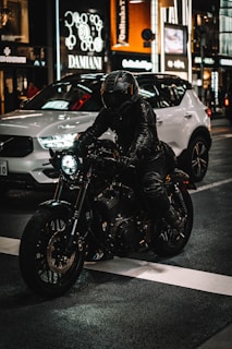 A person wearing a black leather jacket and helmet rides a motorcycle in a city street at night. Bright storefront lights illuminate the background, and a white car is parked nearby, enhancing the urban setting.