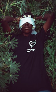A person lies peacefully on their back surrounded by lush green leaves. Their eyes are covered with a cloth adorned with small white flowers, and they wear a black shirt with a heart and crossbones design.