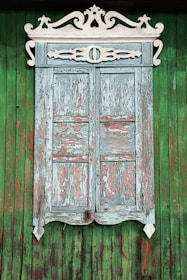 A rustic wooden window with intricate white carvings adorning the top edge. The window shutters are weathered, with peeling light blue and pinkish paint. The background consists of vertical green wooden planks, creating a vivid contrast with the age-worn window.