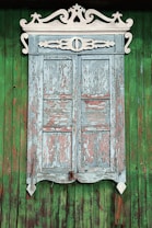 A rustic wooden window with intricate white carvings adorning the top edge. The window shutters are weathered, with peeling light blue and pinkish paint. The background consists of vertical green wooden planks, creating a vivid contrast with the age-worn window.