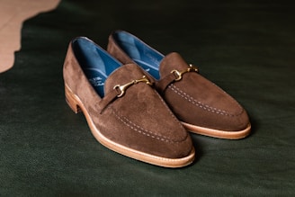 A pair of brown suede loafers with a gold bit detail on the front, resting on a dark green textured surface. The shoes have a wooden sole and a rounded toe design. The interior lining is blue, providing a contrasting color to the brown exterior.
