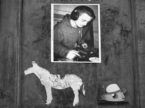 A monochrome collage featuring a photograph of a person wearing headphones, possibly a DJ. Underneath the photo, there are two pieces of paper art stuck on a textured wooden surface. The left figure resembles a horse silhouette with intricate patterns, and the right is a cartoonish character wearing a cap.
