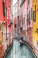 A gondola navigates a narrow canal flanked by tall, colorful buildings with red and yellow walls. The architecture is characterized by brickwork and shuttered windows, creating a charming and historic atmosphere.