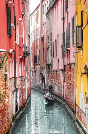 A gondola navigates a narrow canal flanked by tall, colorful buildings with red and yellow walls. The architecture is characterized by brickwork and shuttered windows, creating a charming and historic atmosphere.