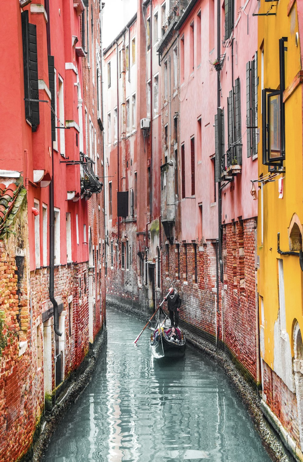 uomo in giacca nera che cavalca sulla barca sul fiume fra gli edifici di cemento rosso e giallo durante